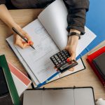 Pessoa analisa documentos financeiros sobre uma mesa, usando calculadora ao lado de um computador.
