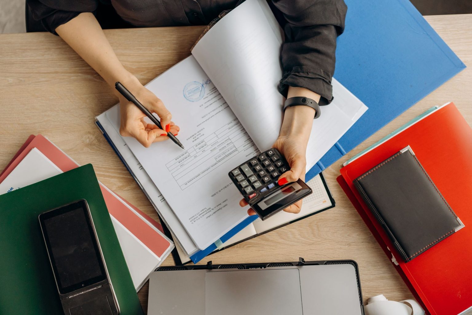 Pessoa analisa documentos financeiros sobre uma mesa, usando calculadora ao lado de um computador.